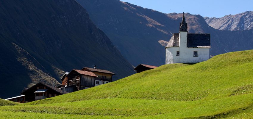 Kapelle in Vrin rechts im Bild auf einer satt grünene Wiese. Im Hintergrund das Dorf und Berge.
