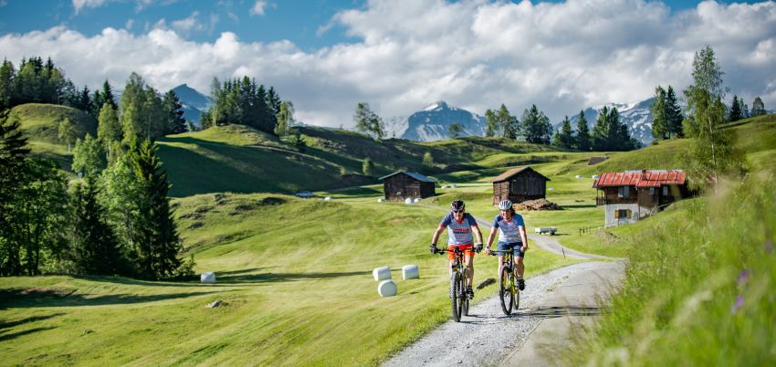 Zwei Biker fahren auf einem Kiesweg. Im Hintergrund geschnittene Wiese mit Heuballen drauf. Rundherum einige Bäume und ein Stall und ein Berghaus.