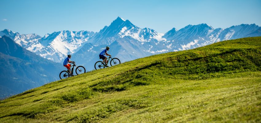 Im Vordergrund des Bildes ist eine Wiese. Im HIntergrund sind schneebedeckte Berge und zwei Biker fahre die Wiese hoch.